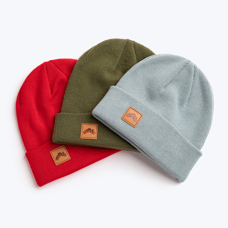 A trio flat lay shot of the ALDRI SUR recycled acrylic beanie in Spice Red, recycled acrylic beanie in Moss Green, and cotton acrylic blend in soft Glacier Blue. All have custom leather logo patch squares in the bottom left corner of the fold